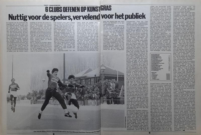Spelers in actie tijdens een voetbalwedstrijd op kunstgras, met publiek op de achtergrond.