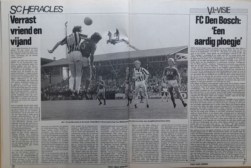 Voetbalwedstrijd met spelers en scheidsrechter op het veld, met publiek op de achtergrond.